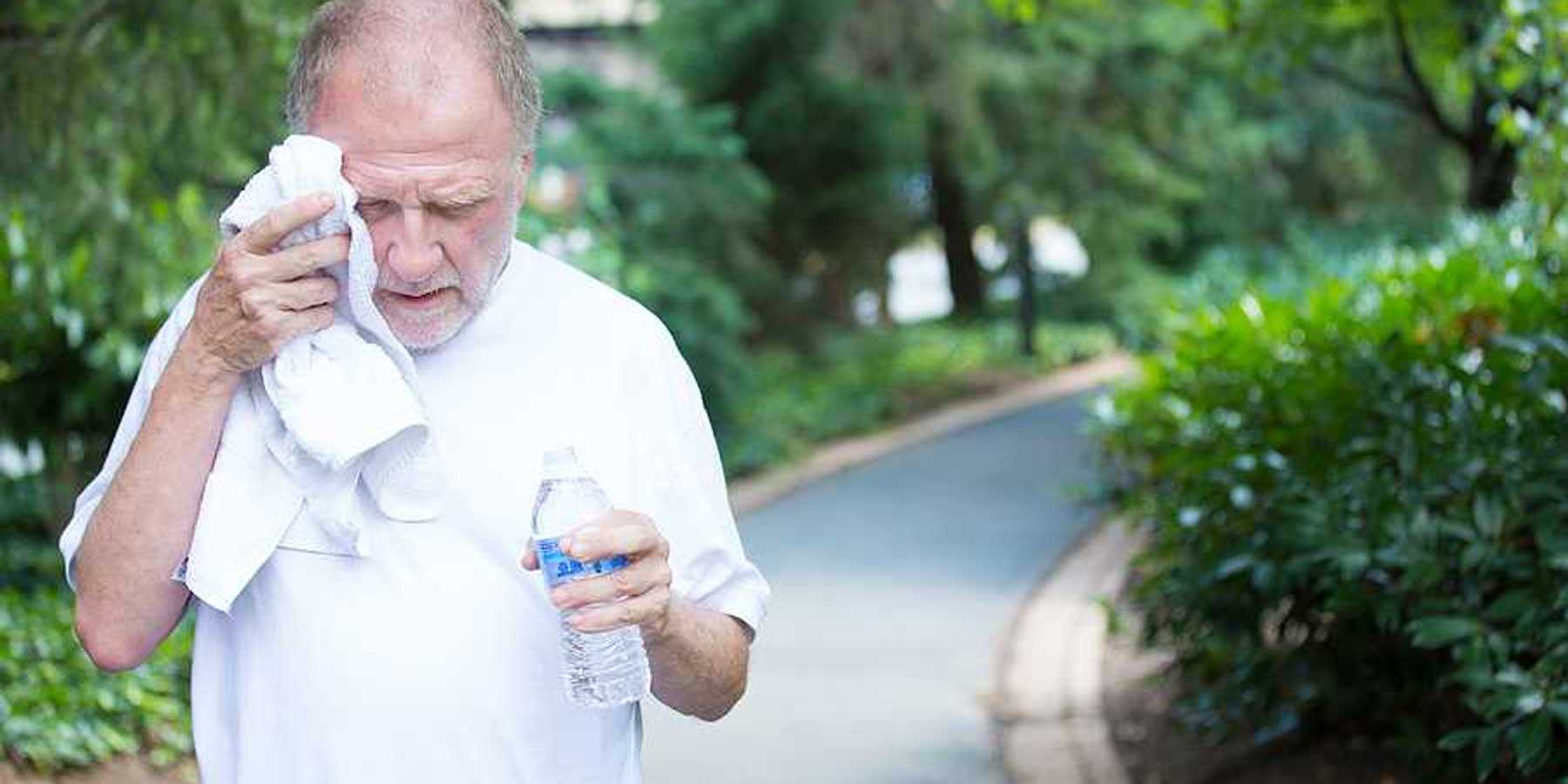 Closeup portrait old gentleman in white shirt having difficulties with extreme heat high temperature wiping sweat from face very tired isolated green trees paved road background
