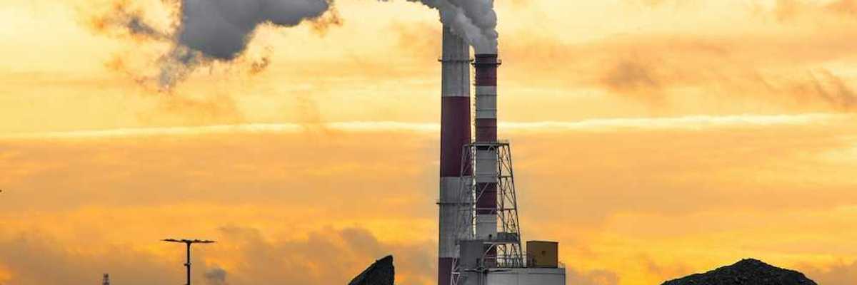 Coal fired power plant with piles of coal beneath red & white stacks emitting smoke