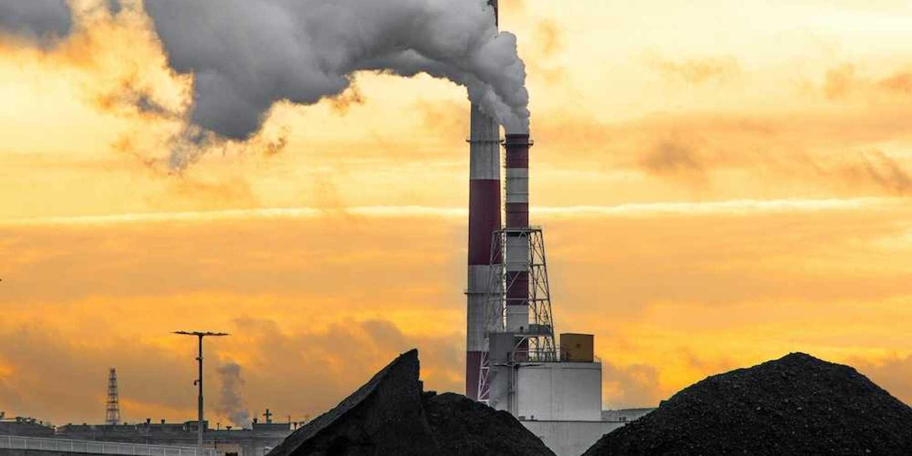 Coal fired power plant with piles of coal beneath red & white stacks emitting smoke