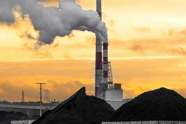 Coal fired power plant with piles of coal beneath red & white stacks emitting smoke