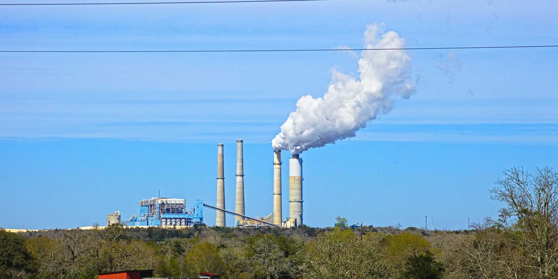 Coal fired power plant with smoke stacks spewing on a hill and a green field in foreground.