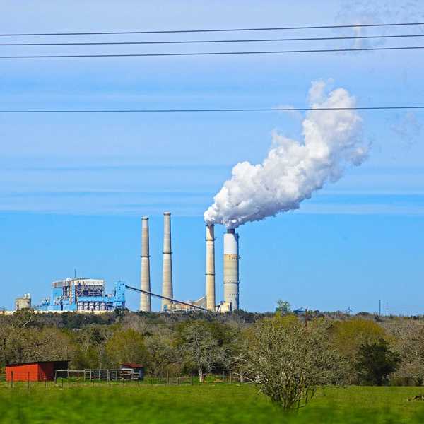 Coal fired power plant with smoke stacks spewing on a hill and a green field in foreground.