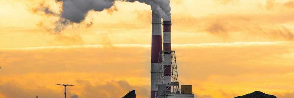 Coal plant emitting smoke from red and white smokestacks adjacent to large piles of coal and coal ash.