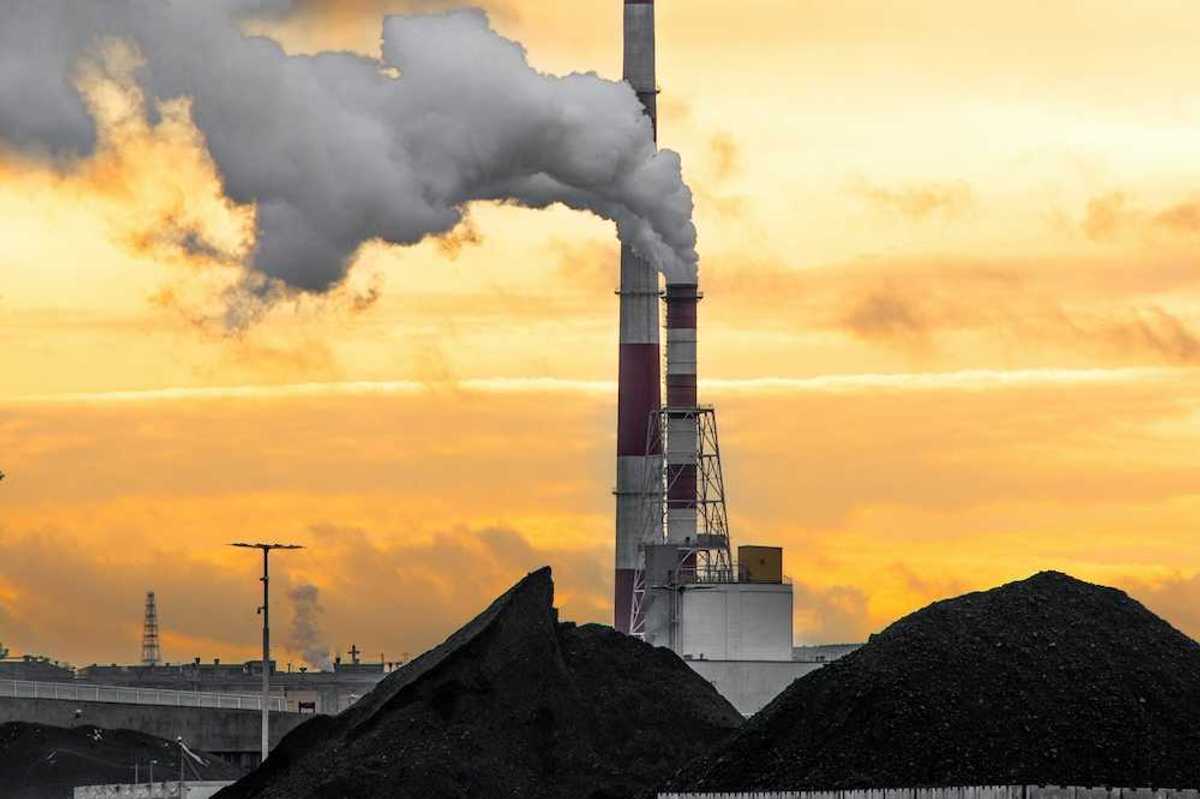 Coal plant emitting smoke from red and white smokestacks adjacent to large piles of coal and coal ash.