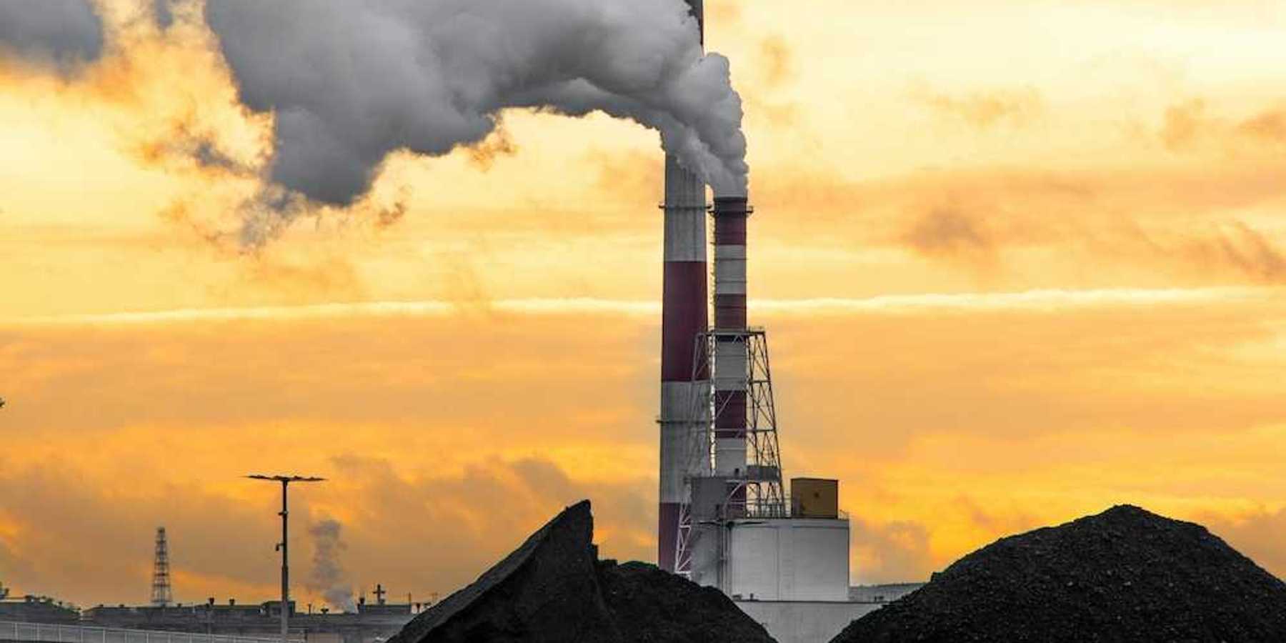 Coal plant emitting smoke from red and white smokestacks adjacent to large piles of coal and coal ash.