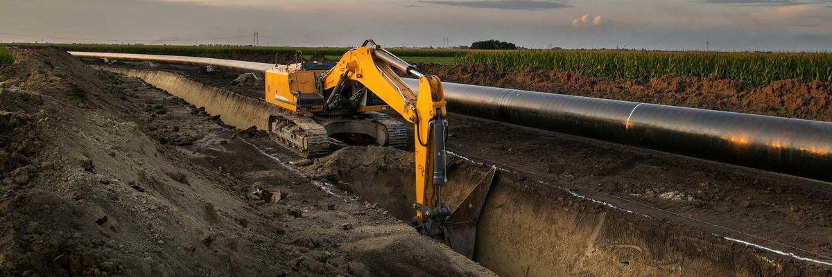 Construction equipment digs a trench along an oil pipeline in a field.