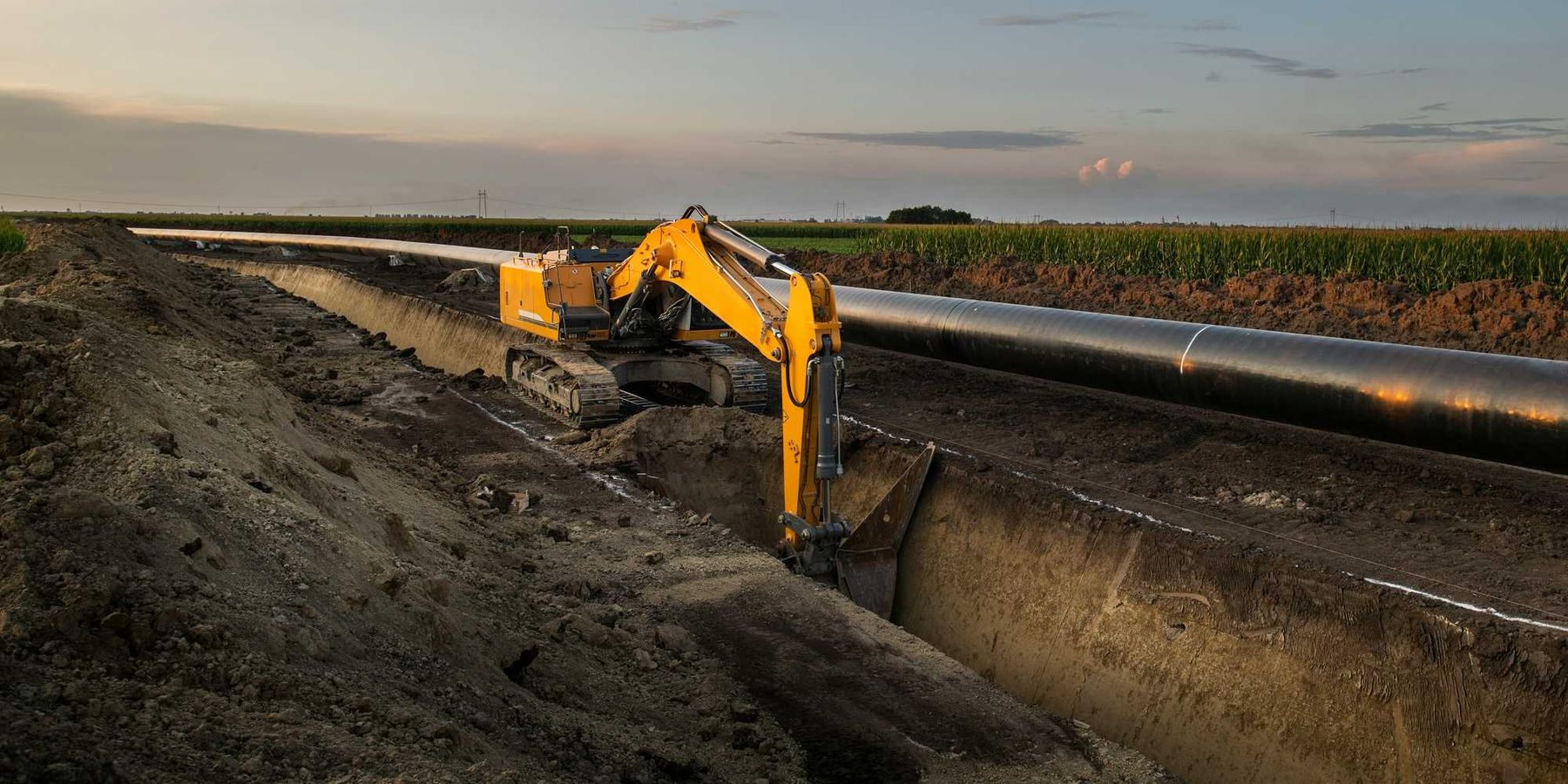 Construction equipment digs a trench along an oil pipeline in a field.