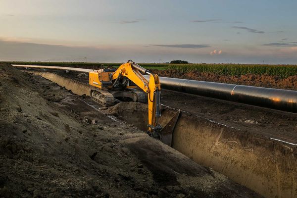 Construction equipment digs a trench along an oil pipeline in a field.