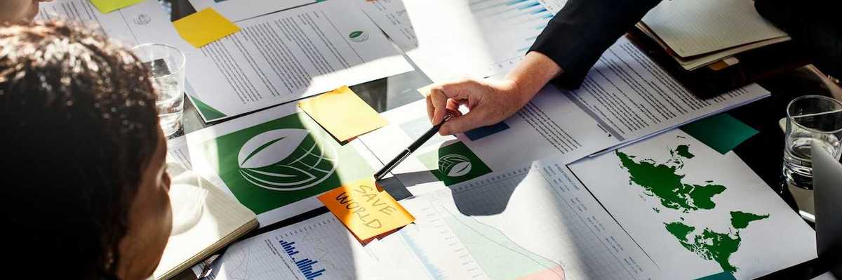 Corporate types gathered around desk full of paperwork with post-it note in center reading "SAVE WORLD."