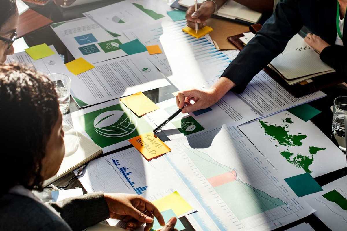 Corporate types gathered around desk full of paperwork with post-it note in center reading "SAVE WORLD."