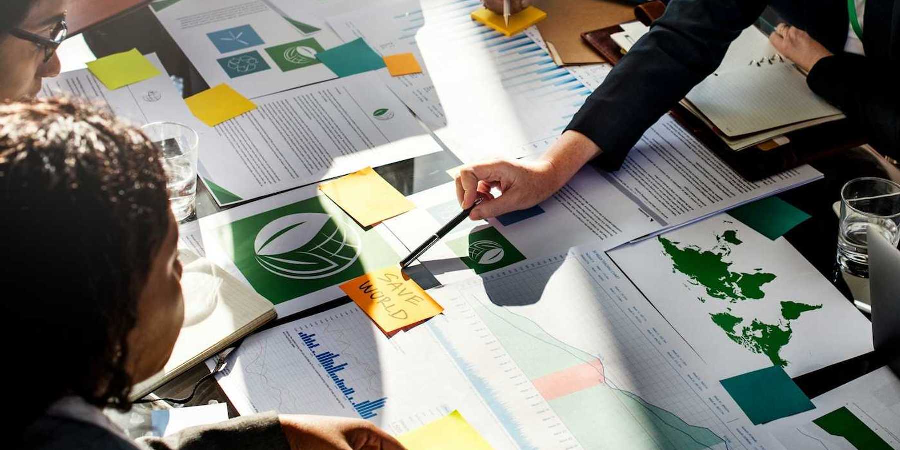 Corporate types gathered around desk full of paperwork with post-it note in center reading "SAVE WORLD."