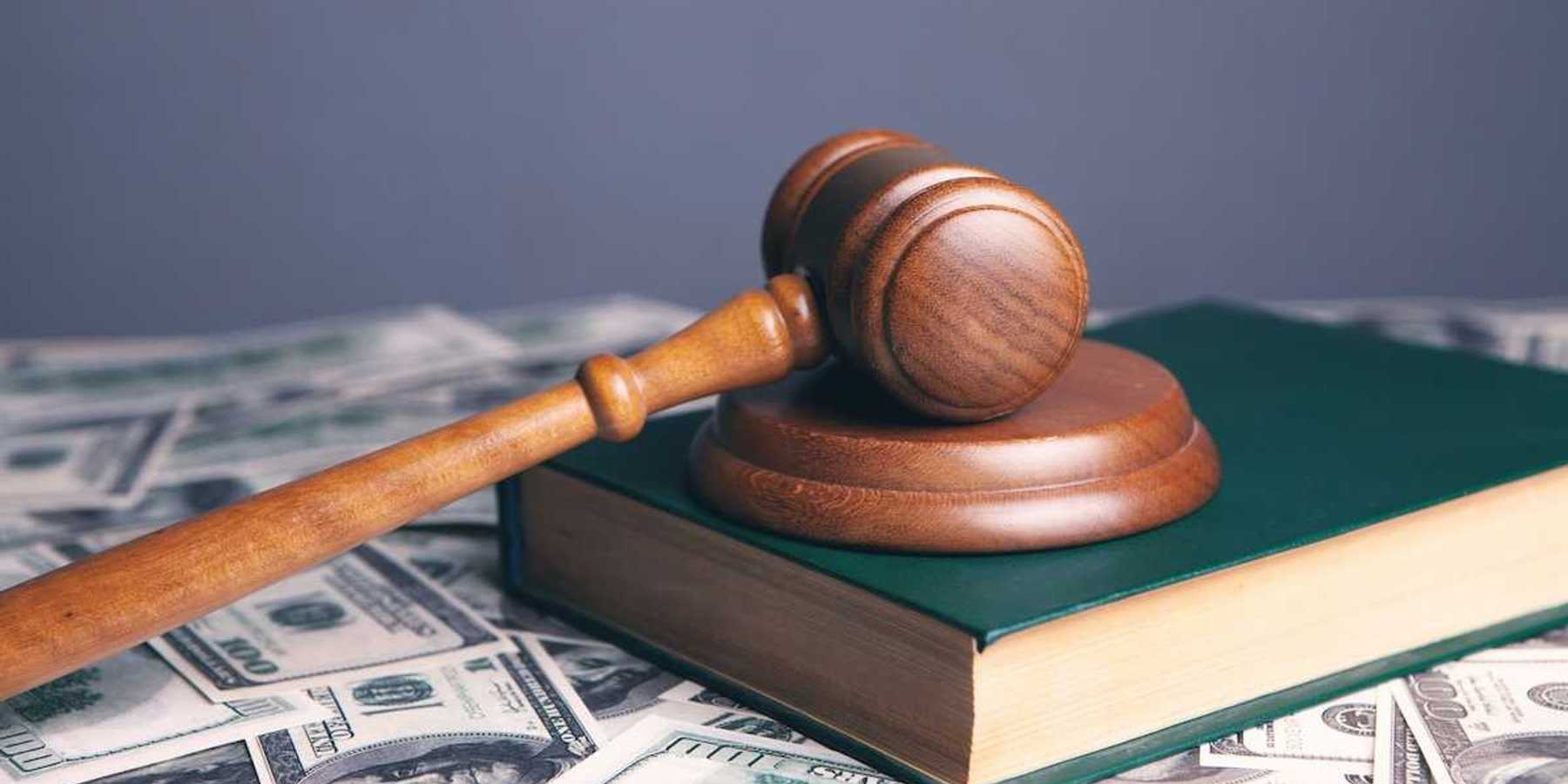 Court gavel resting on unnamed book which is sitting on scattered $100 bills.
