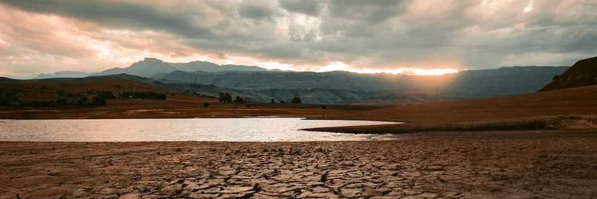 Cracked drought-ravaged foreground, receding water level of lake in background, sun rising or setting on distant hills