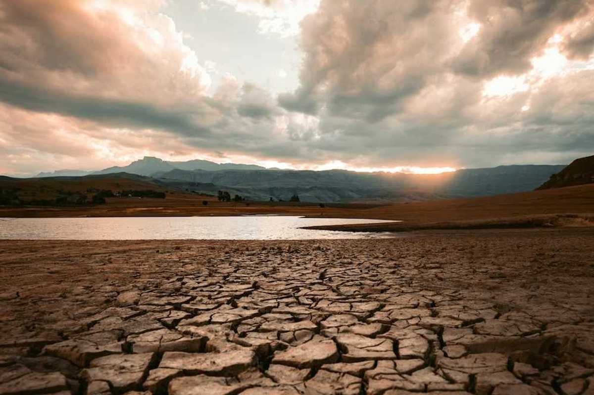 Cracked drought-ravaged foreground, receding water level of lake in background, sun rising or setting on distant hills