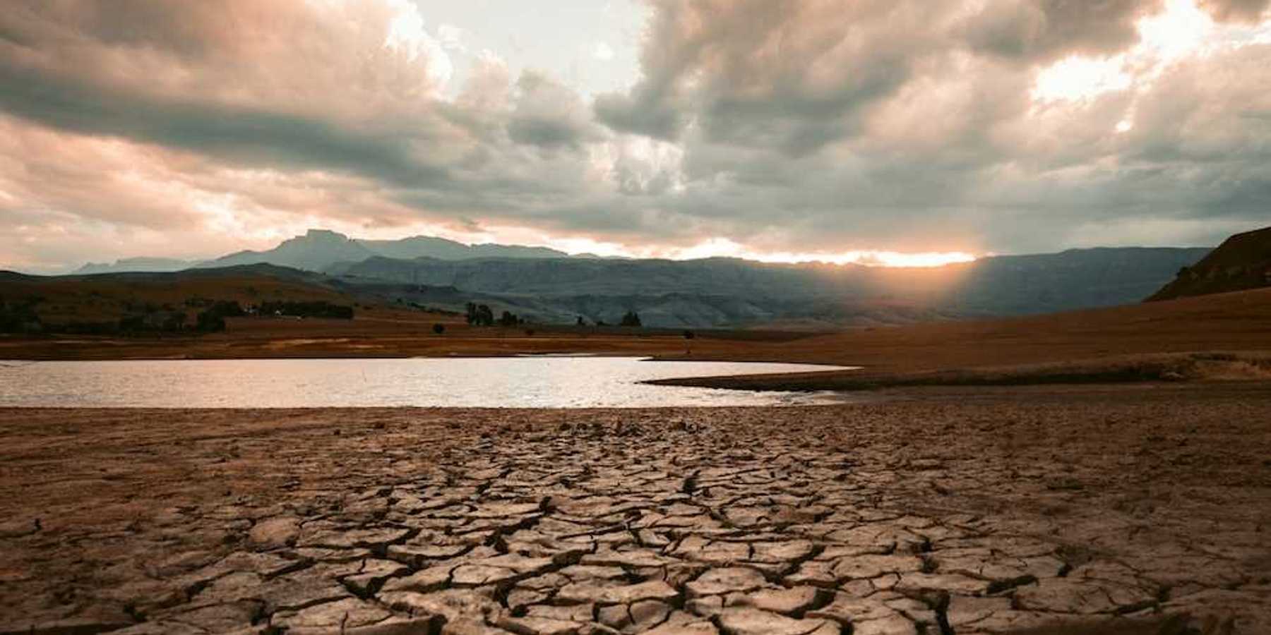 Cracked drought-ravaged foreground, receding water level of lake in background, sun rising or setting on distant hills
