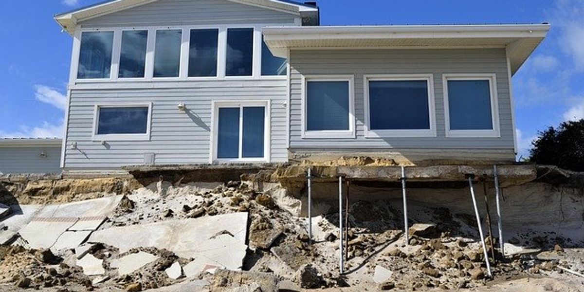 Damaged beach house with erosion visible at base and metal stilts supporting part of the structure.