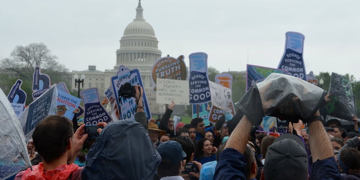 Demonstrators hold signs supporting science on a rainy day, with the US Capitol building in background.