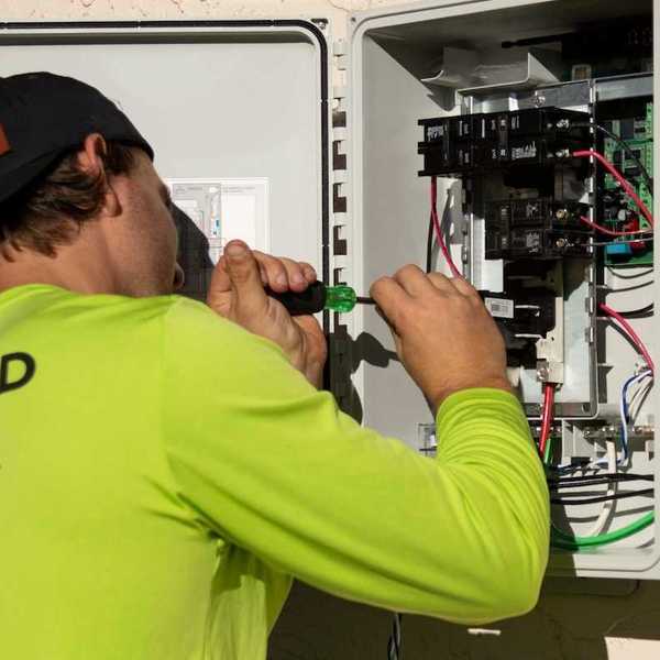 Electrician in yellow-green shirt using a screwdriver while working on an electrical service panel