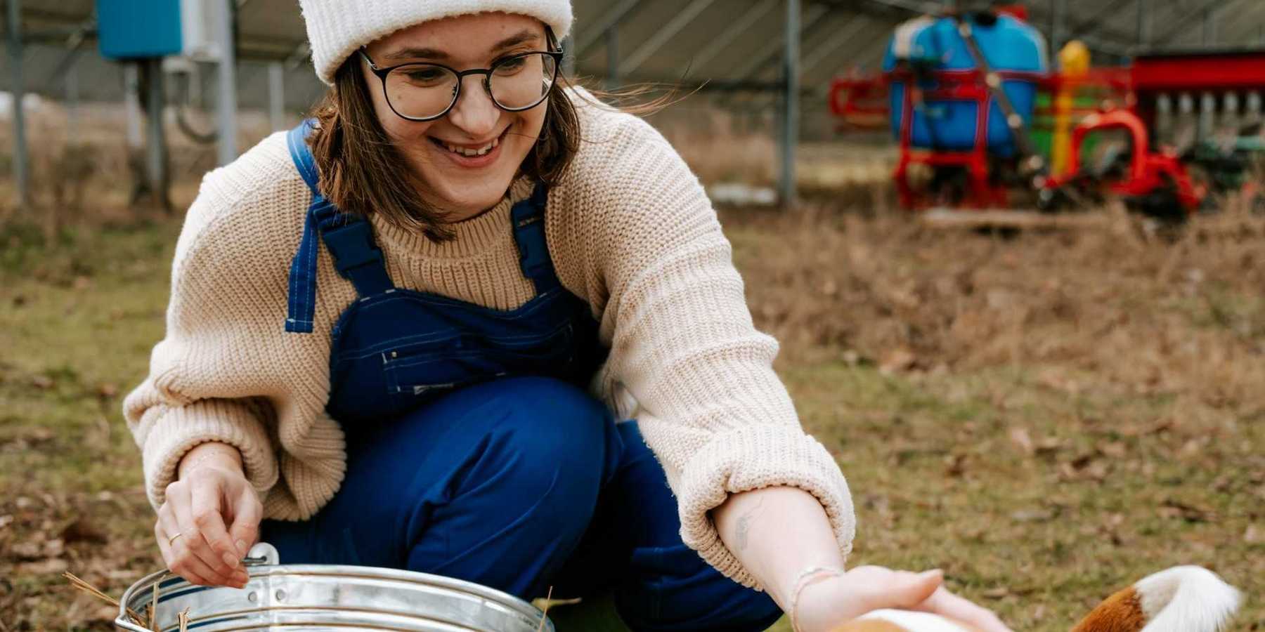 Farmer with stainless steel bucket squatting behind solar panels and petting a brown and white dog