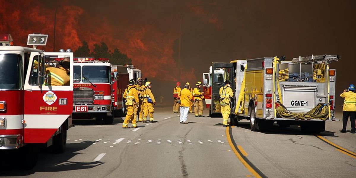 Firetrucks and firefighters gathered on a highway with a raging wildfire in the background.