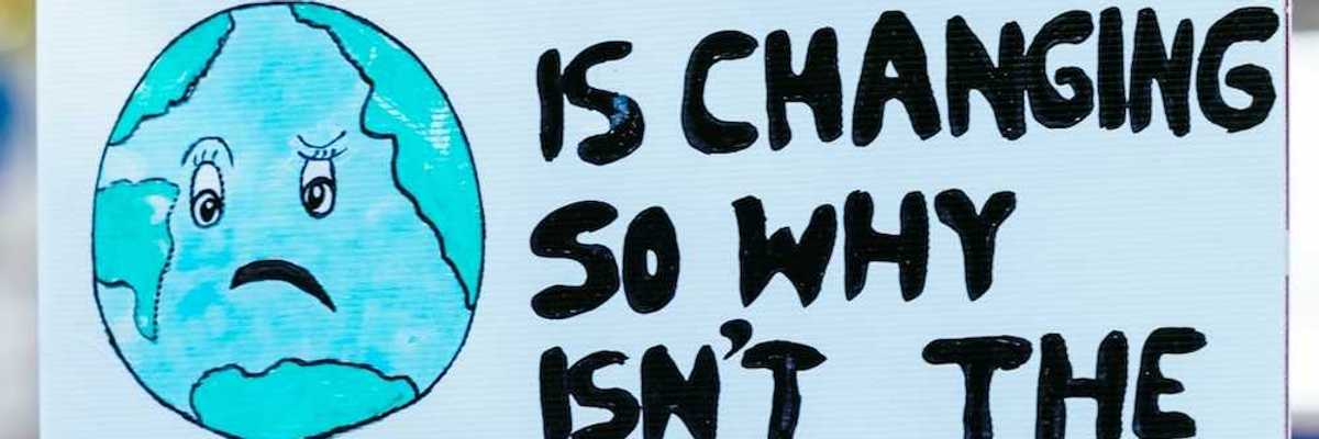 Hand holding handmade sign that reads "THE CLIMATE IS CHANGING SO WHY ISN'T THE SYSTEM?"