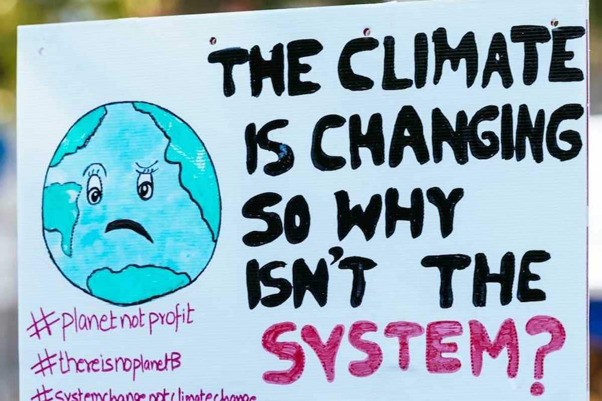 Hand holding handmade sign that reads "THE CLIMATE IS CHANGING SO WHY ISN'T THE SYSTEM?"