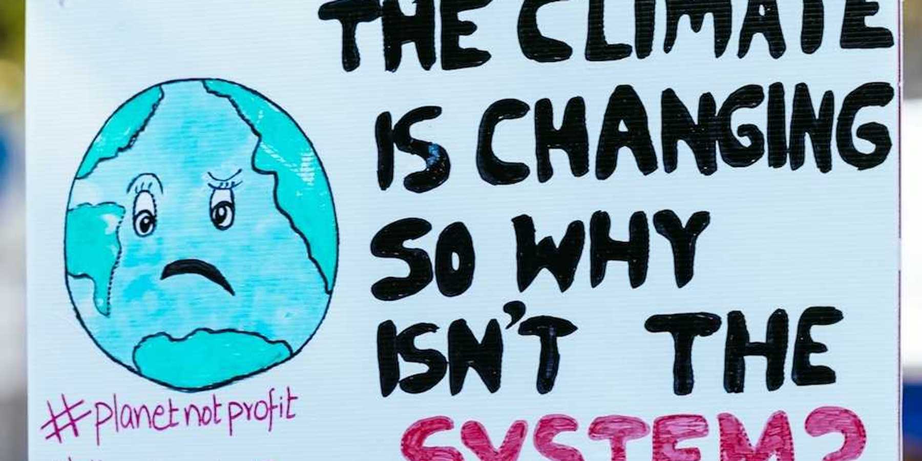 Hand holding handmade sign that reads "THE CLIMATE IS CHANGING SO WHY ISN'T THE SYSTEM?"