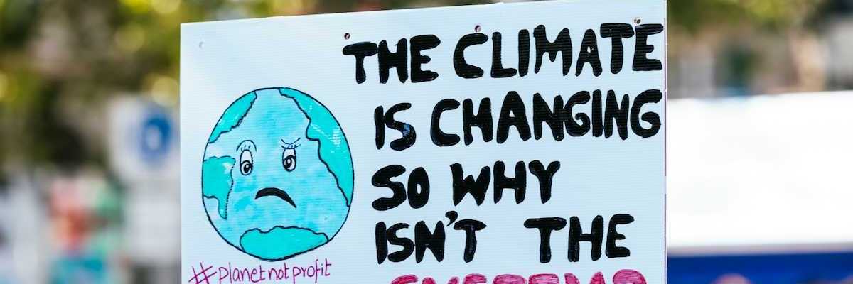 Hand holding sign: "THE CLIMATE IS CHANGING SO WHY ISN'T THE SYSTEM?"