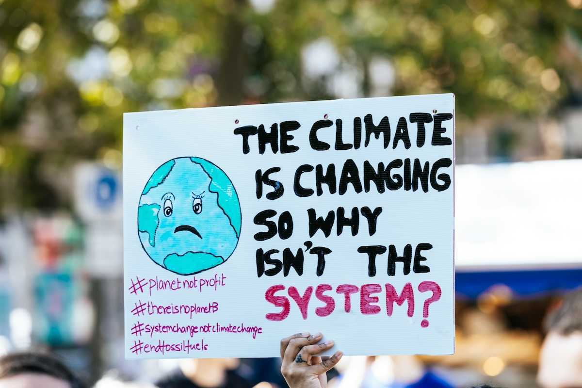 Hand holding sign: "THE CLIMATE IS CHANGING SO WHY ISN'T THE SYSTEM?"