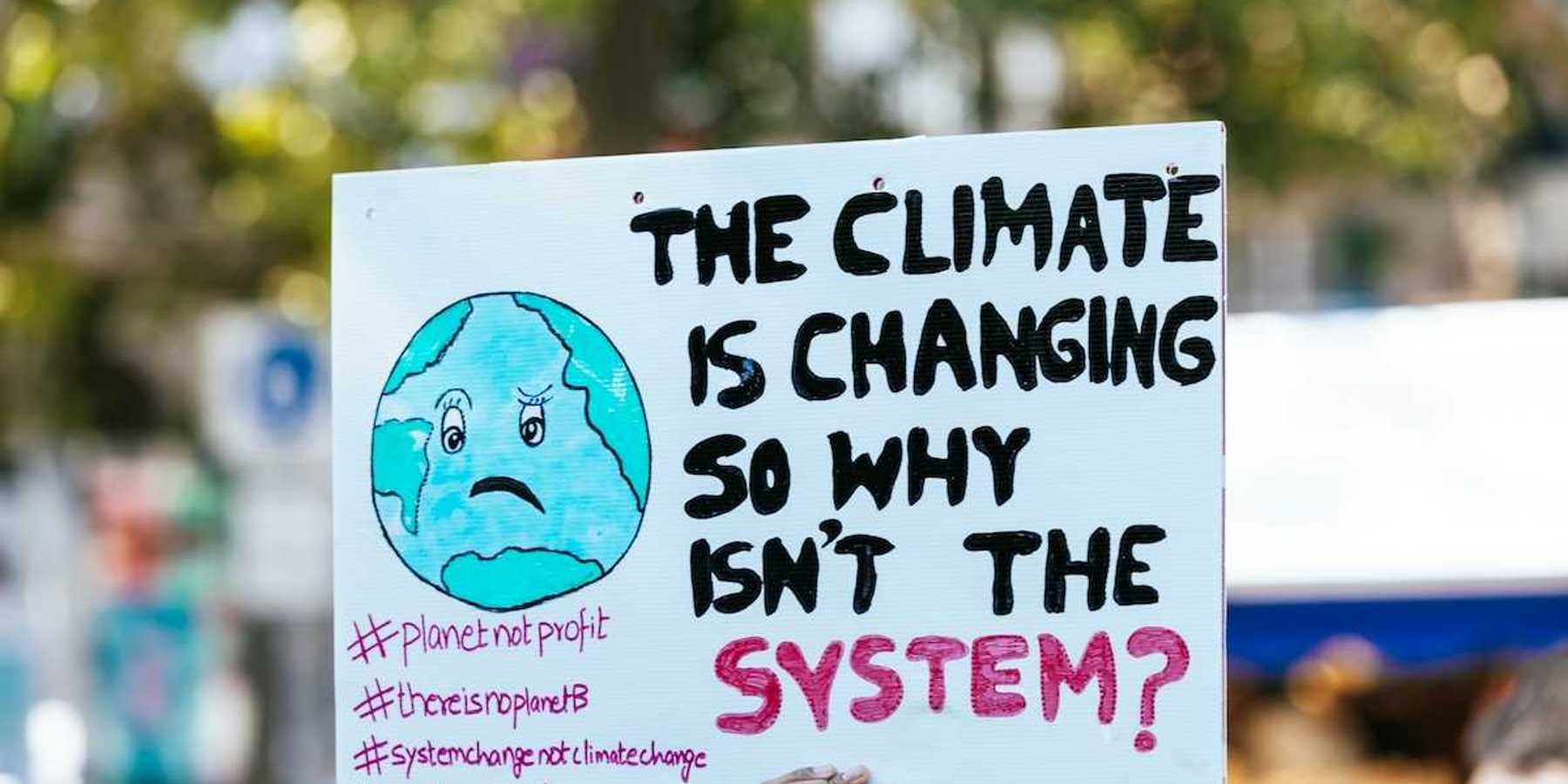 Hand holding sign: "THE CLIMATE IS CHANGING SO WHY ISN'T THE SYSTEM?"