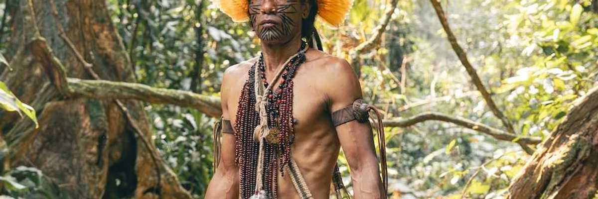 Indigenous Amazonian clothed in traditional dress, carrying traditional implements & weapons