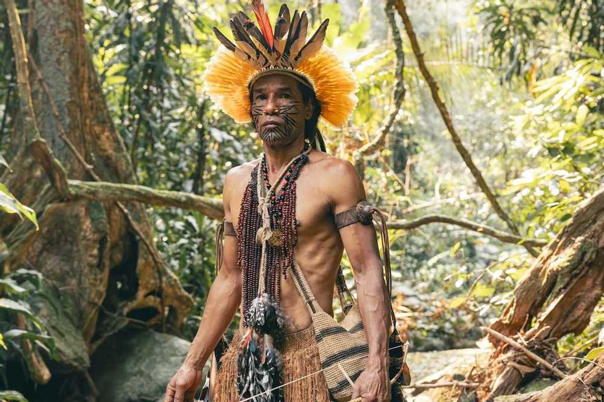 Indigenous Amazonian clothed in traditional dress, carrying traditional implements & weapons