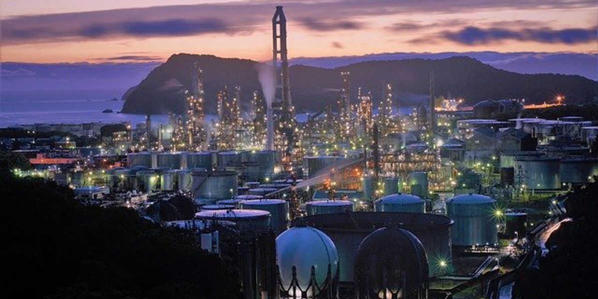 Industrial buildings and smokestacks on the coast with mountains and sunset in the background.