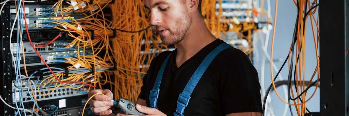 Male technician dressed in black t-shirt and blue bibbed overalls testing a rack of electronic gear