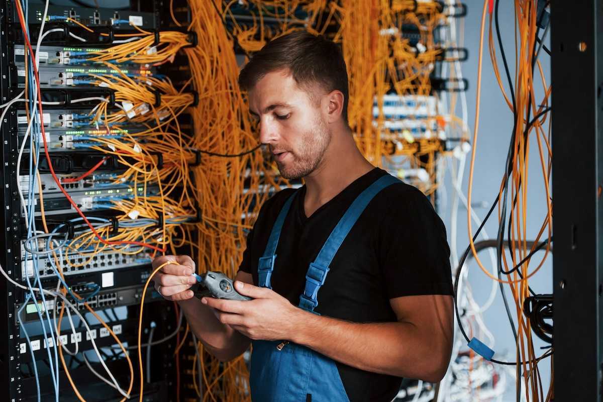 Male technician dressed in black t-shirt and blue bibbed overalls testing a rack of electronic gear