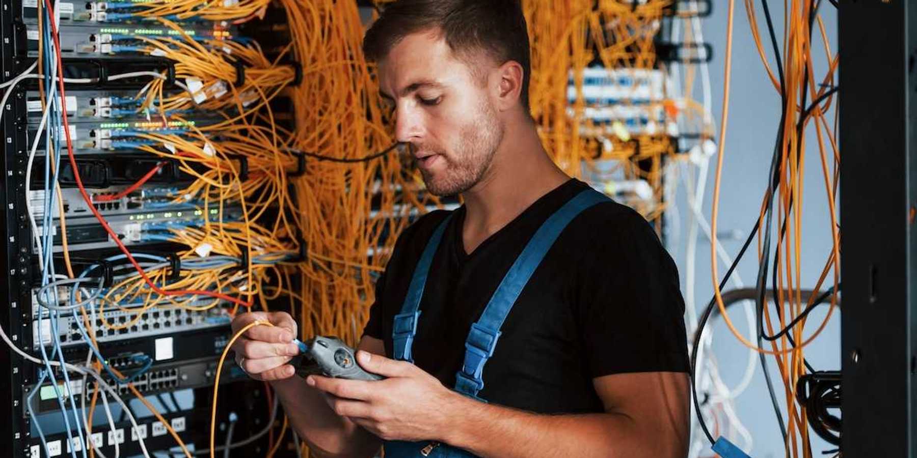 Male technician dressed in black t-shirt and blue bibbed overalls testing a rack of electronic gear