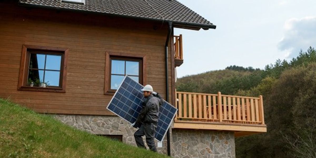 Man carrying a solar panel up a grass-covered slope near a home.