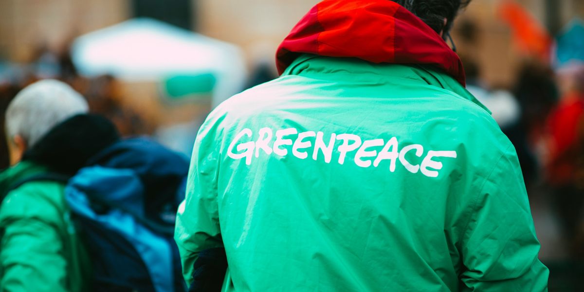 Man in green jacket with the word Greenpeace on the back walking on street during daytime.