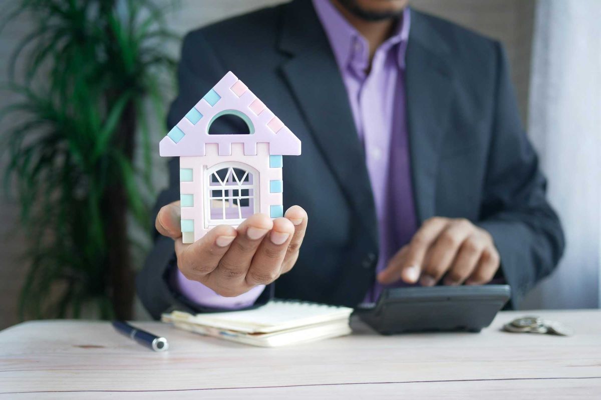 man in suit holding a toy model of a house and using a calculator