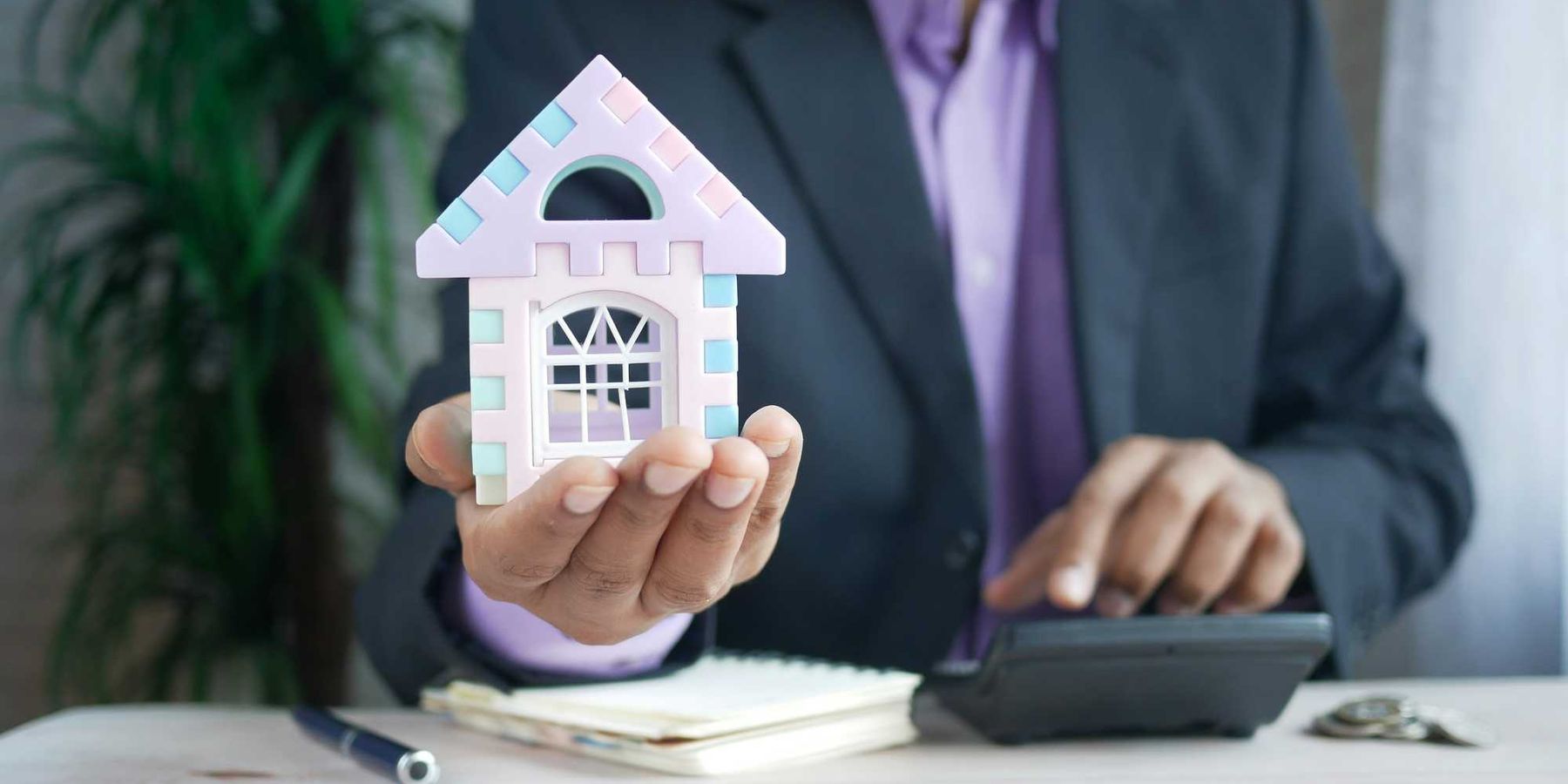 man in suit holding a toy model of a house and using a calculator