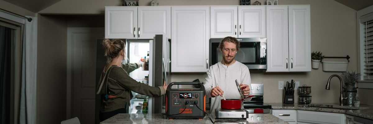 Man prepares dinner in kitchen while woman opens refrigerator during power outage while appliances are powered by a portable power station.