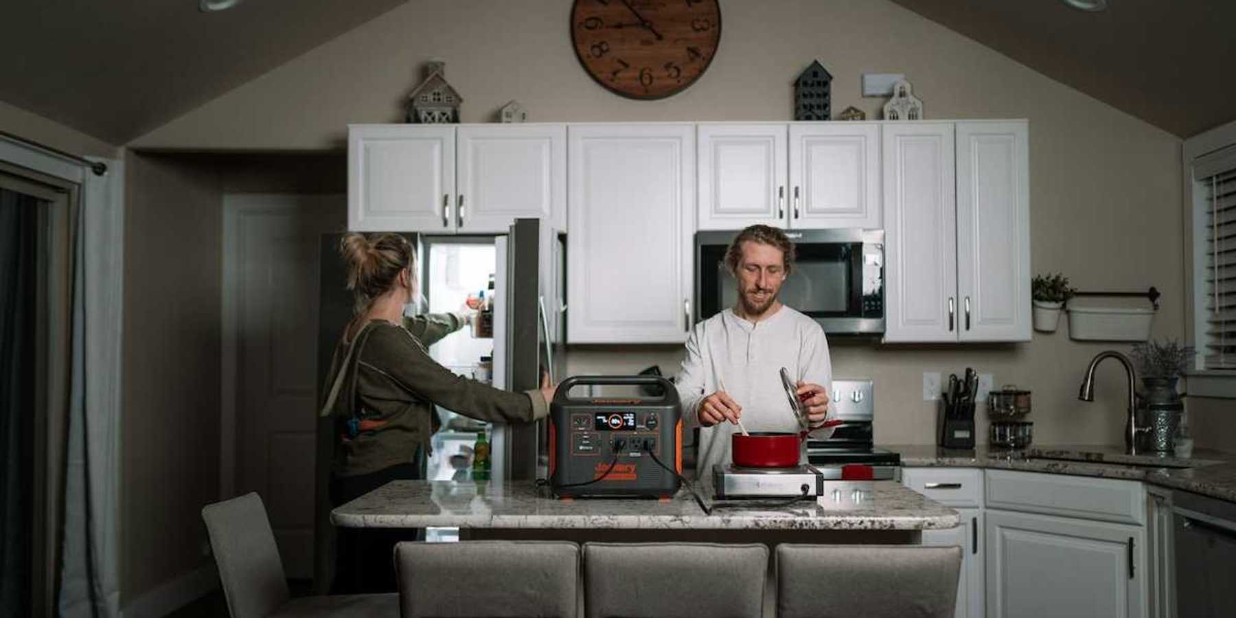 Man prepares dinner in kitchen while woman opens refrigerator during power outage while appliances are powered by a portable power station.