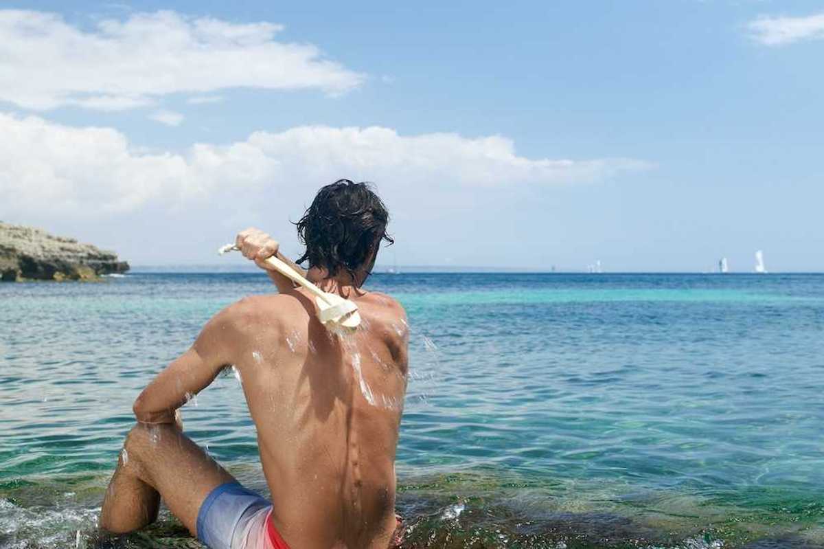 Man scrubbing his own back with a long-handled brush while sitting in Mediterranean Sea water.