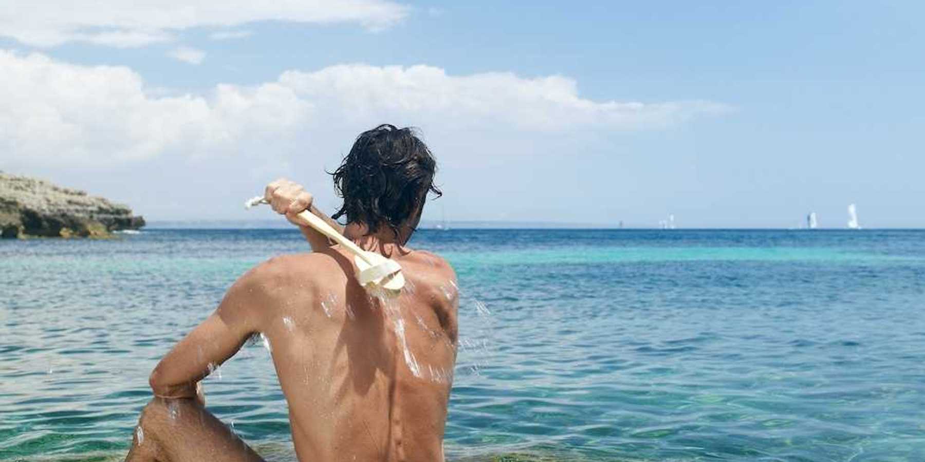 Man scrubbing his own back with a long-handled brush while sitting in Mediterranean Sea water.