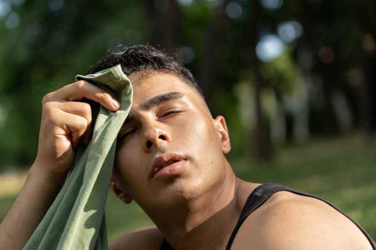 Man wiping the sweat off of his forehead with a shirt while sitting outside in a park