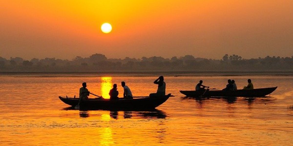Men silhouetted in boats on a river with the sun setting in the background and reflecting orange on the water.