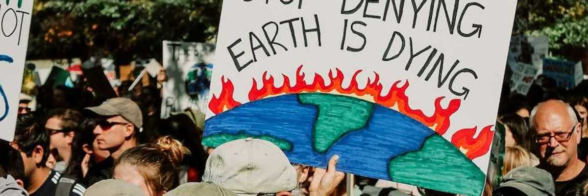Mixed crowd taking part in climate protest prominently featuring a sign reading, "Stop Denying Earth is Dying"