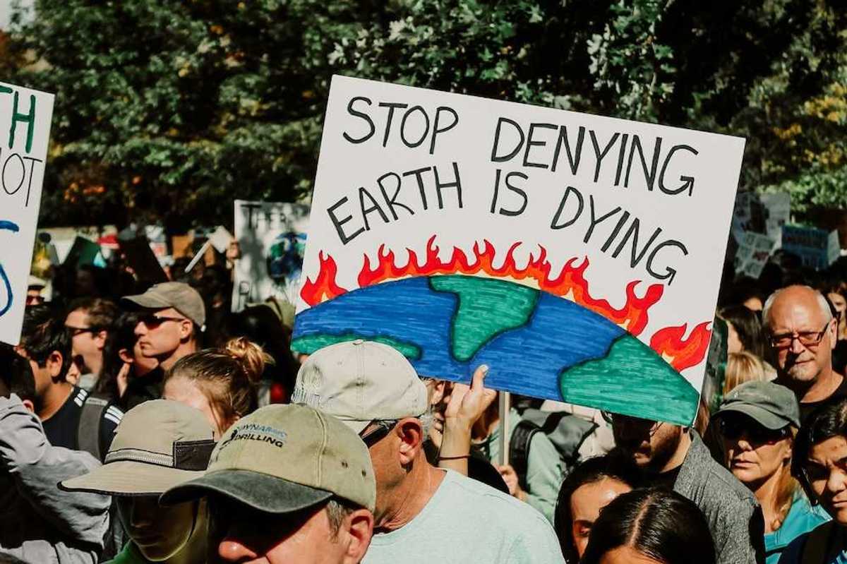 Mixed crowd taking part in climate protest prominently featuring a sign reading, "Stop Denying Earth is Dying"
