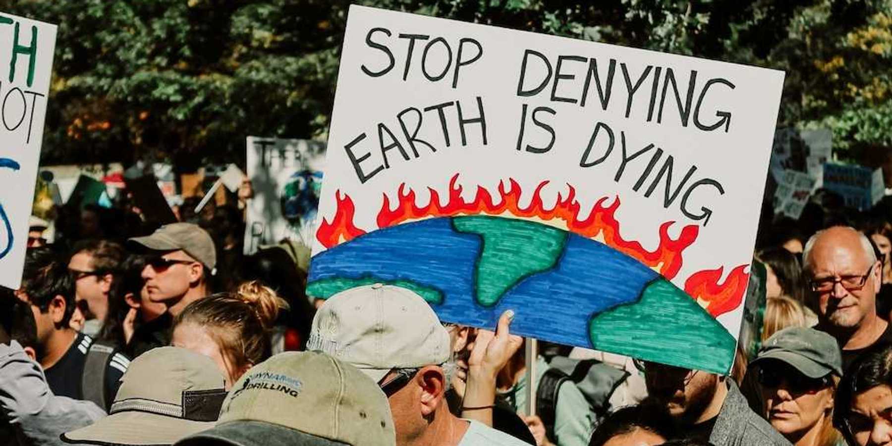 Mixed crowd taking part in climate protest prominently featuring a sign reading, "Stop Denying Earth is Dying"