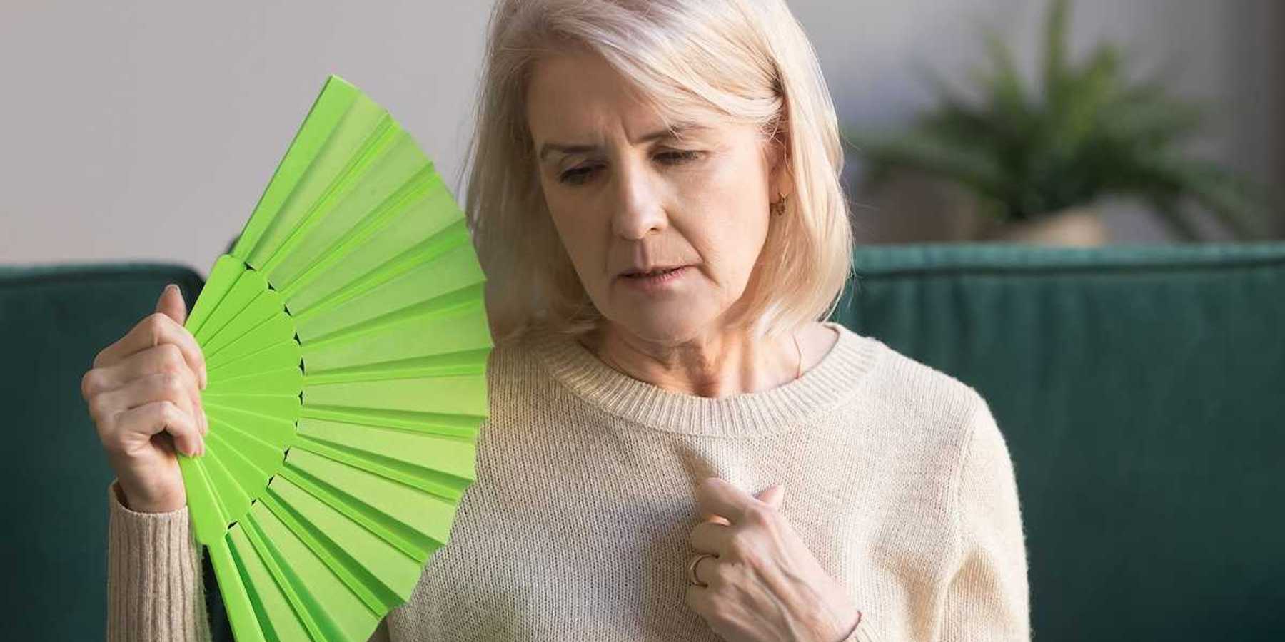 Overheated elderly woman sitting on couch waving green fan.